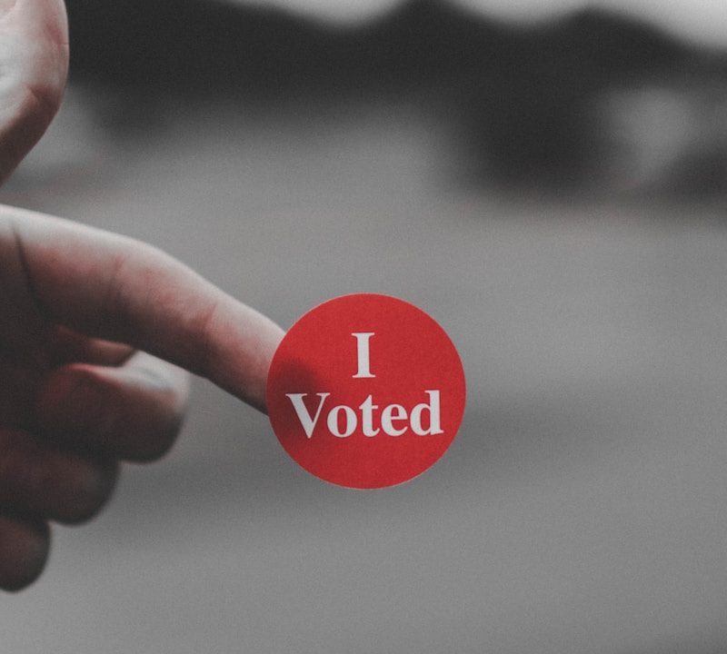 a hand holding a red button that says i vote